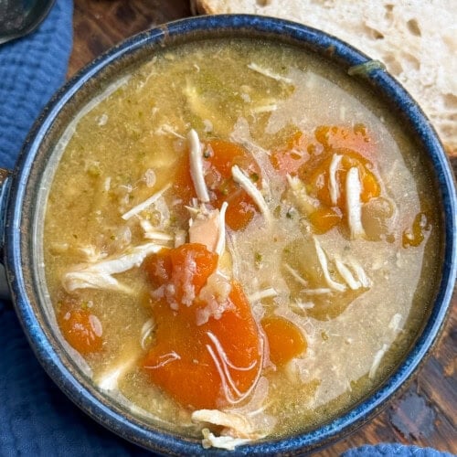 A bowl of chicken soup with shredded chicken, carrot slices, and broth sits on a blue cloth, with slices of rustic bread in the background.