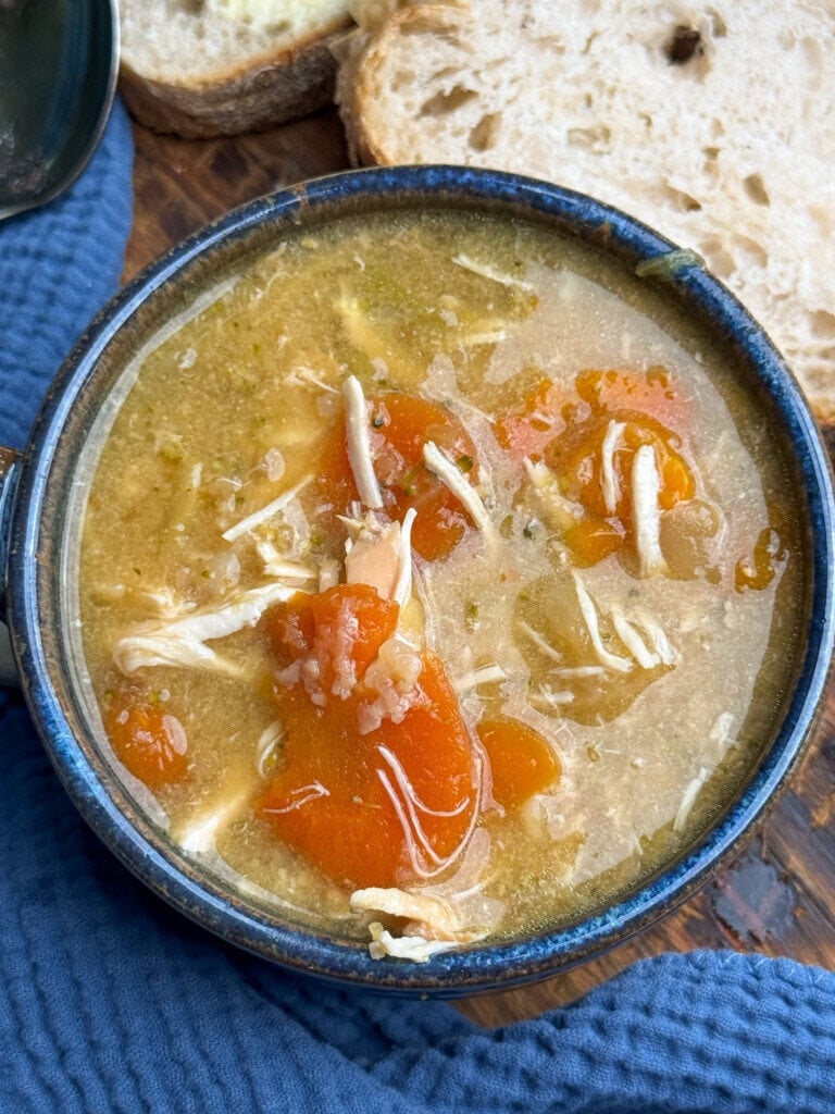 A bowl of chicken soup with shredded chicken, carrot slices, and broth sits on a blue cloth, with slices of rustic bread in the background.