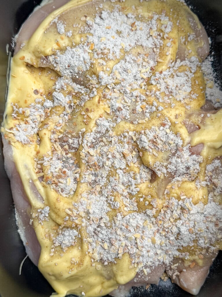 Raw chicken breasts coated with a layer of mustard, sprinkled with flour and sesame seeds, placed in a dark baking dish ready to be cooked.