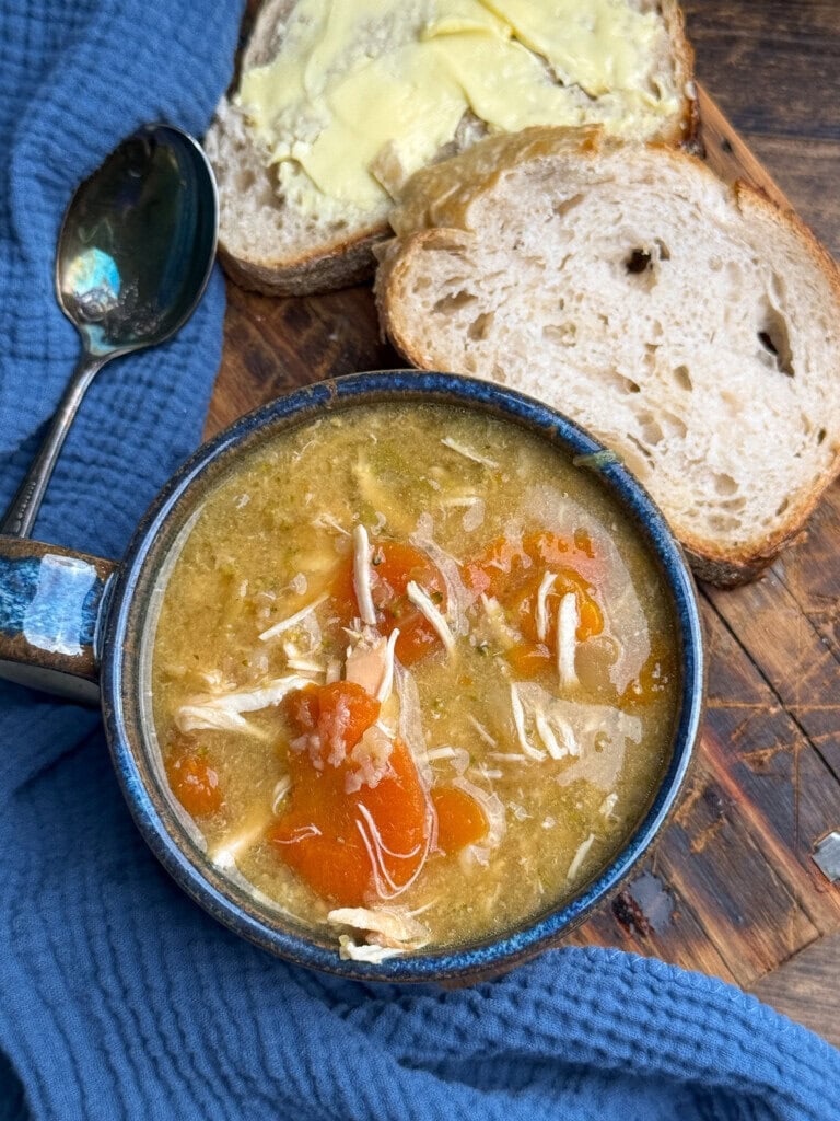 A bowl of chicken and vegetable soup with chunks of carrot, beside two slices of buttered bread on a wooden surface, with a spoon and blue cloth nearby.