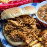 A pulled chicken sandwich on a bun is served with crinkle-cut fries and baked beans on a blue-and-white plate. A basket of fries is visible in the background.