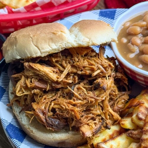 A pulled chicken sandwich on a bun is served with crinkle-cut fries and baked beans on a blue-and-white plate. A basket of fries is visible in the background.