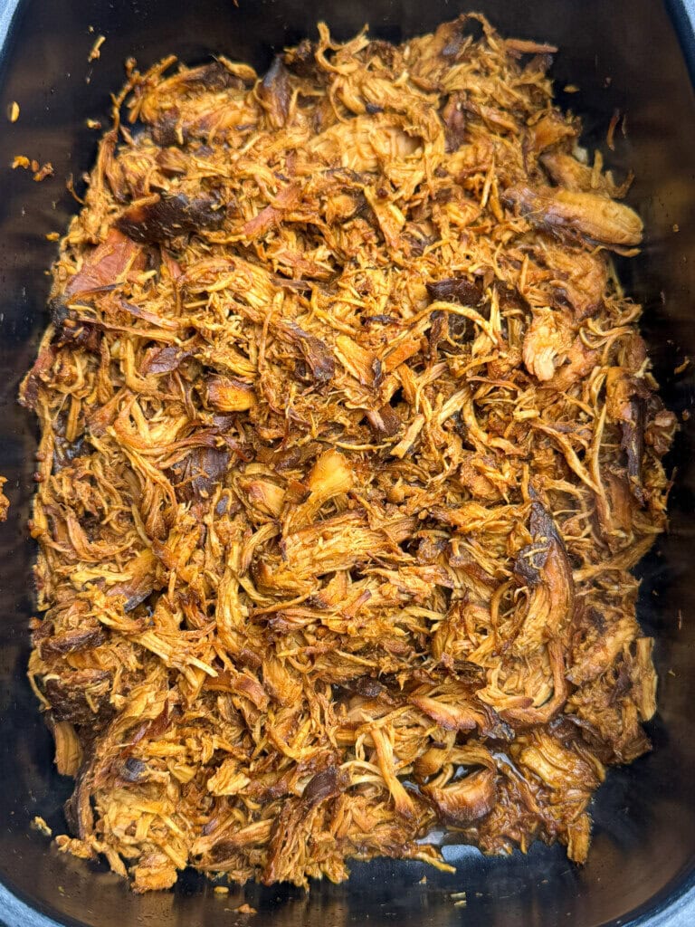 A close-up of shredded, seasoned cooked chicken in a black container, showcasing its tender texture and rich, golden-brown color.