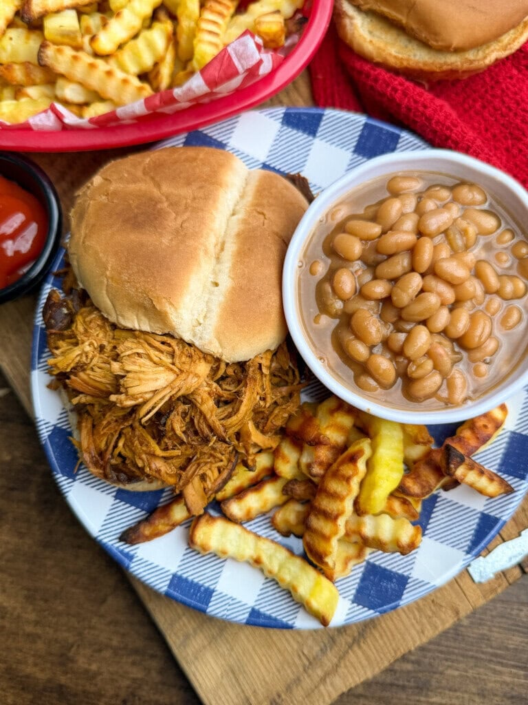 A plate with a pulled chicken sandwich, crinkle-cut fries, and baked beans in a bowl. A red basket with more fries and part of a sandwich are in the background, along with a small dish of ketchup.