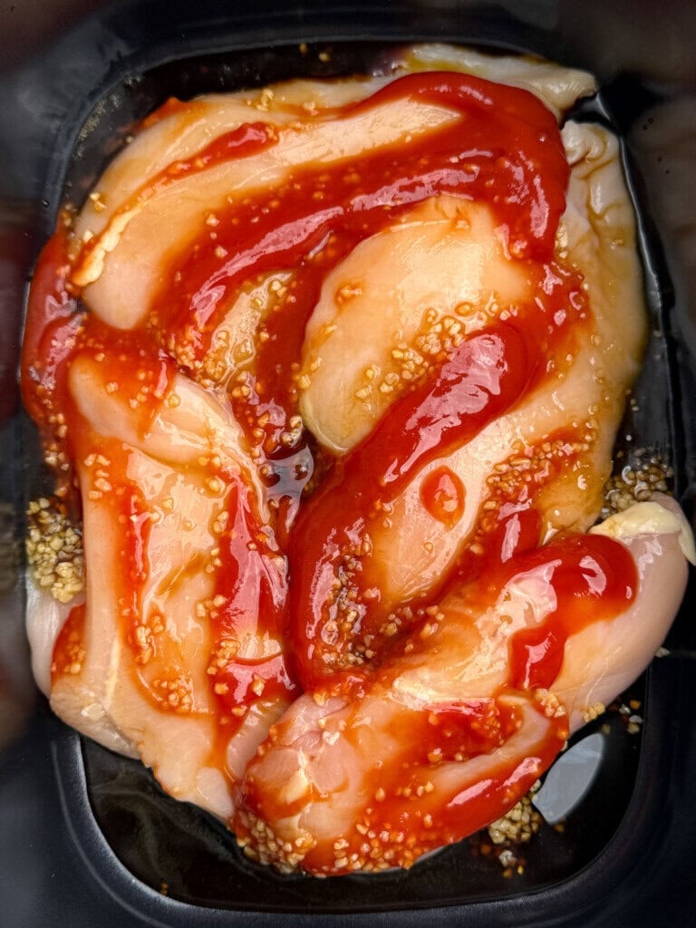 Raw chicken breast strips marinating in a black tray with a mixture of red sauce and visible garlic. The marinade appears glossy and thick, covering most of the chicken pieces.