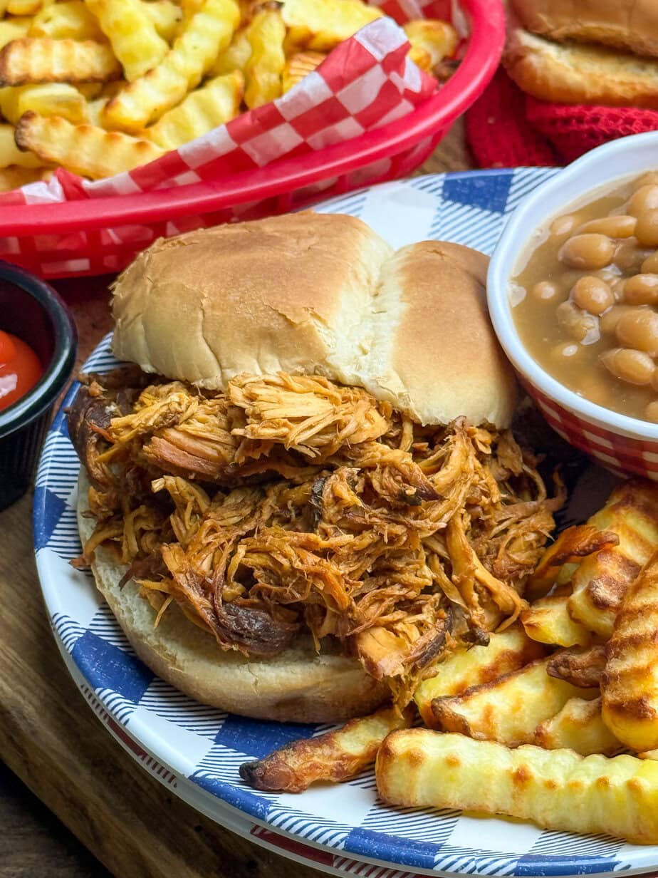 Pulled chicken sandwich on a bun with crinkle-cut fries and baked beans on a blue and white plate. Basket of fries and ketchup are in the background.