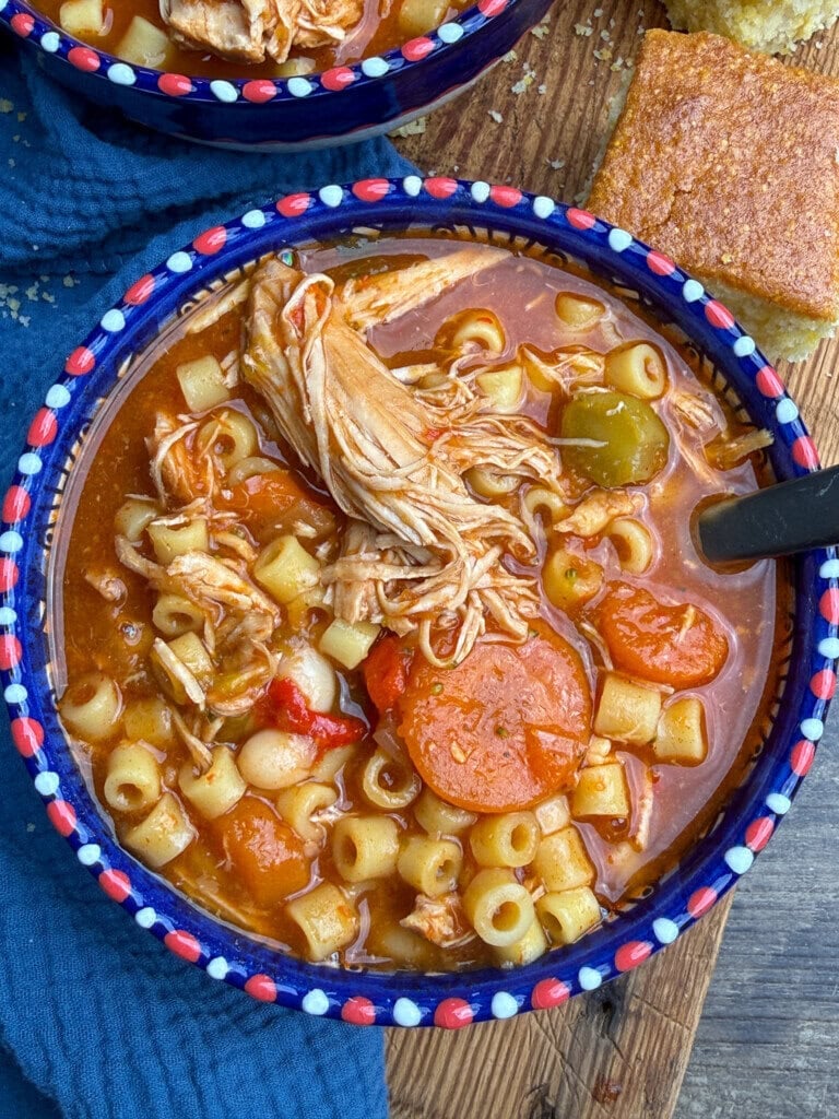 A colorful bowl of chicken noodle soup with ditalini pasta, carrots, bell peppers, and shredded chicken, served with a slice of cornbread on the side.