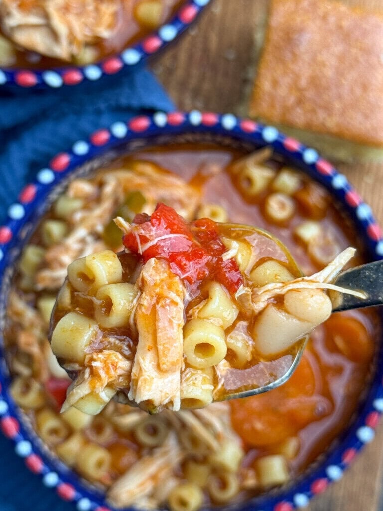 A close-up of a spoon holding pasta, shredded chicken, white beans, and tomatoes over a bowl of soup with a colorful rim.