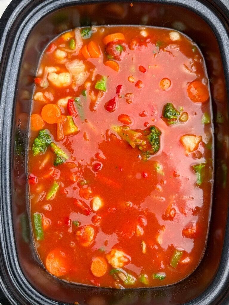 A close-up of vegetable soup in a black container, featuring carrots, broccoli, cauliflower, corn, and red sauce.