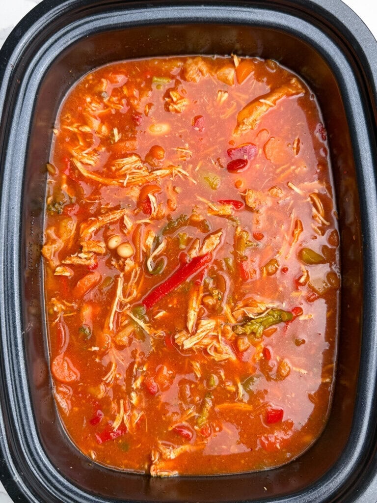 A close-up of a slow cooker filled with chunky chicken chili containing shredded chicken, beans, diced tomatoes, peppers, and a rich red sauce.