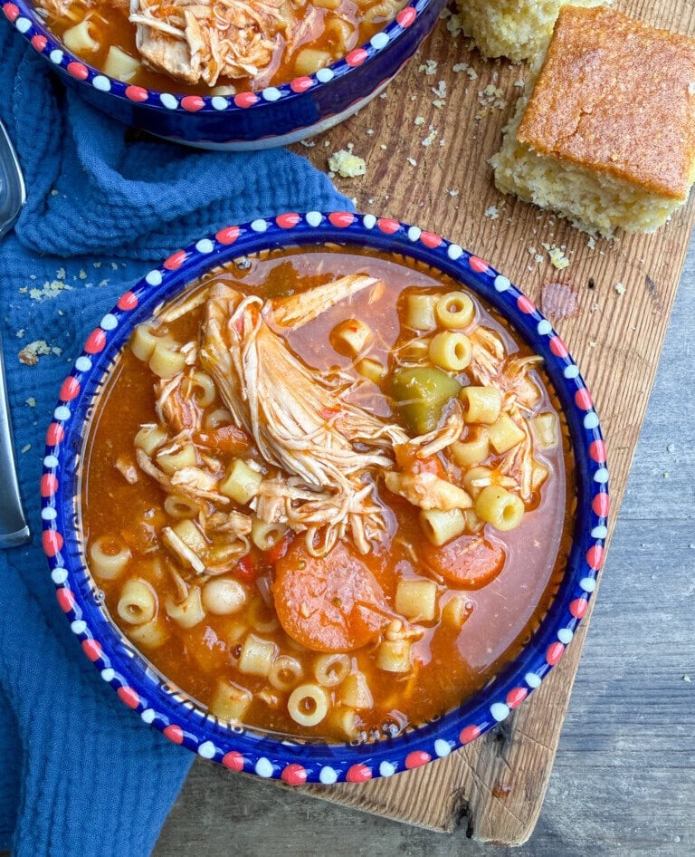 A bowl of chicken noodle soup with ditalini pasta, carrots, and peppers on a wooden board, next to a square piece of cornbread and a blue cloth.