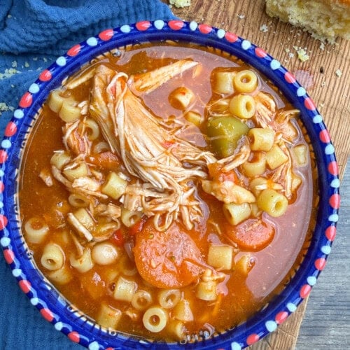 A colorful bowl filled with chicken noodle soup, featuring shredded chicken, ditalini pasta, carrots, celery, and broth, sits on a blue cloth next to a slice of cornbread on a wooden surface.