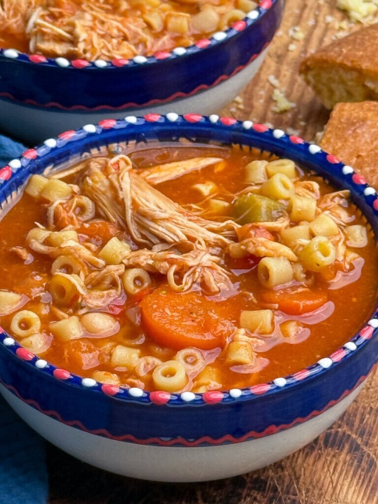 A bowl of chicken noodle soup with ditalini pasta, carrots, and shredded chicken in a tomato-based broth, served on a wooden table with cornbread in the background.