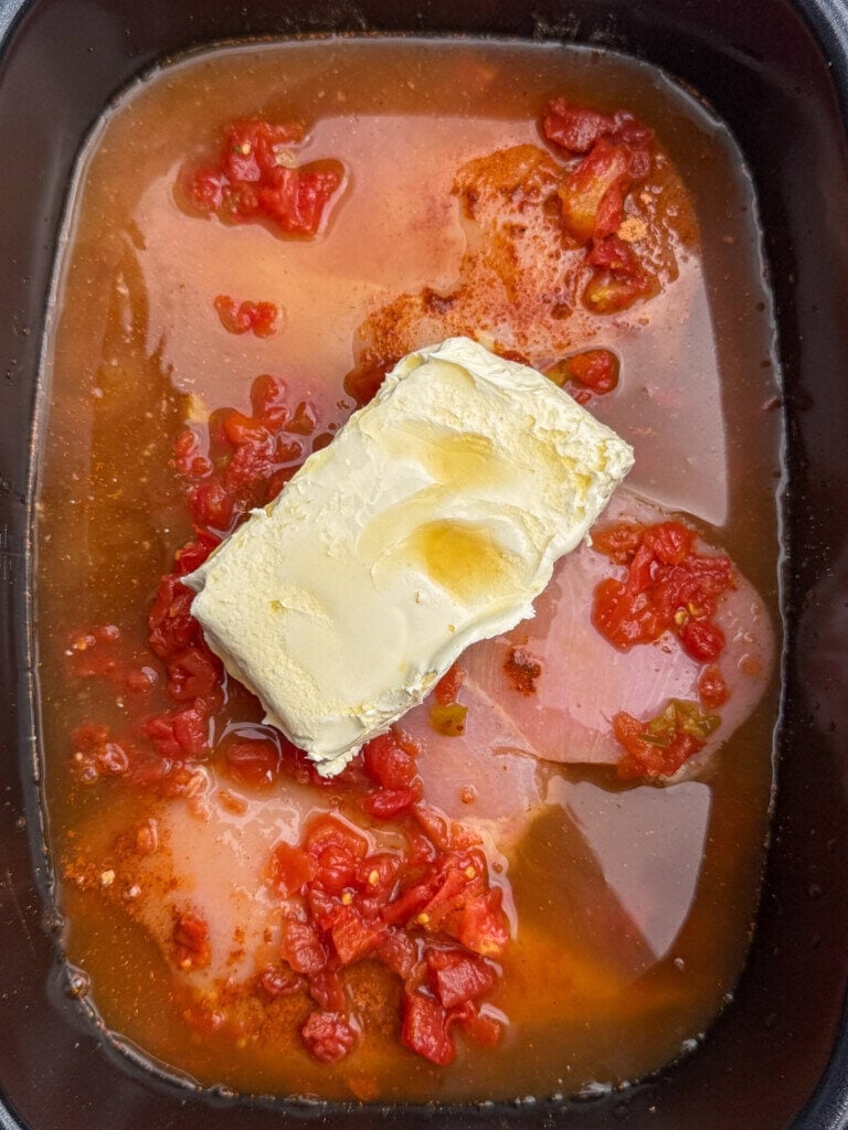 A block of cream cheese sits in a slow cooker with diced tomatoes and liquid, ready to be mixed into a dish.