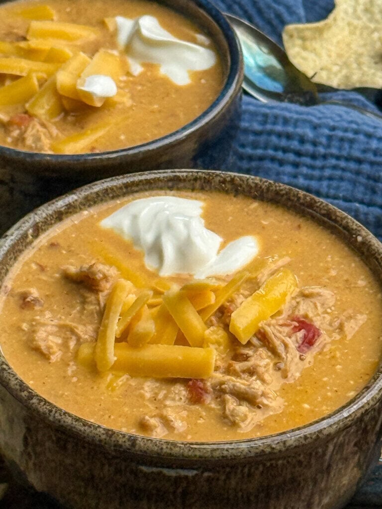 Two bowls of creamy soup topped with shredded cheddar cheese and a dollop of sour cream, placed on a blue cloth with a spoon and tortilla chip in the background.