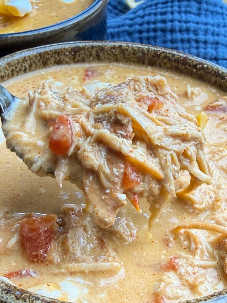 A spoon lifts shredded chicken and tomatoes from a creamy, orange-colored soup in a rustic bowl, with a blue textured cloth in the background.
