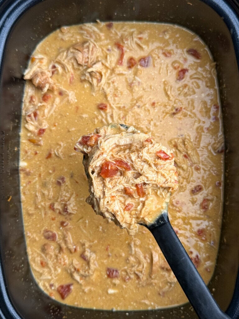 A ladle lifts a serving of creamy shredded chicken with tomatoes from a slow cooker filled with the same dish.