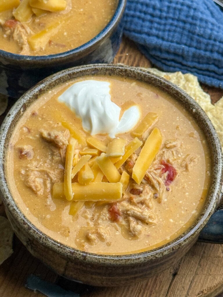 A bowl of creamy chicken soup topped with shredded cheddar cheese and a dollop of sour cream, with another bowl and blue cloth in the background. Tortilla chips are visible on the side.