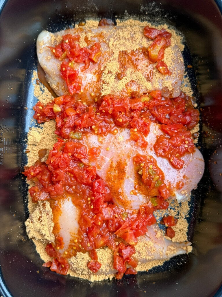 Raw chicken breasts in a baking dish topped with diced tomatoes and sprinkled with a yellowish seasoning.
