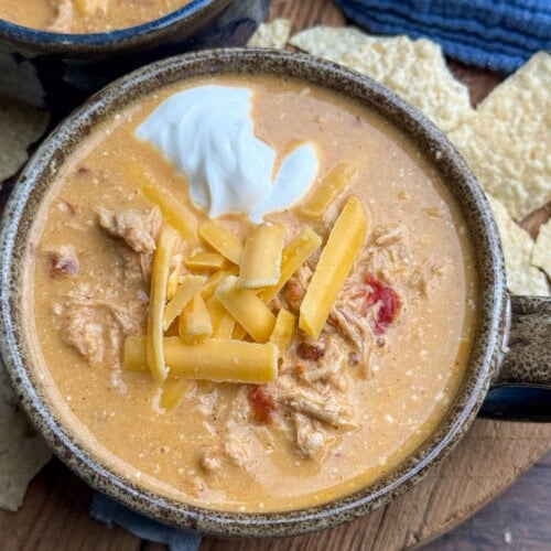 A bowl of creamy chicken soup topped with shredded cheddar cheese and a dollop of sour cream, placed on a wooden surface with tortilla chips and a blue cloth nearby.