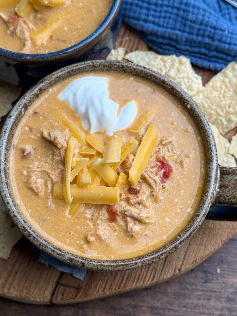 A bowl of creamy chicken soup topped with shredded cheddar cheese and a dollop of sour cream, placed on a wooden surface with tortilla chips and a blue cloth nearby.